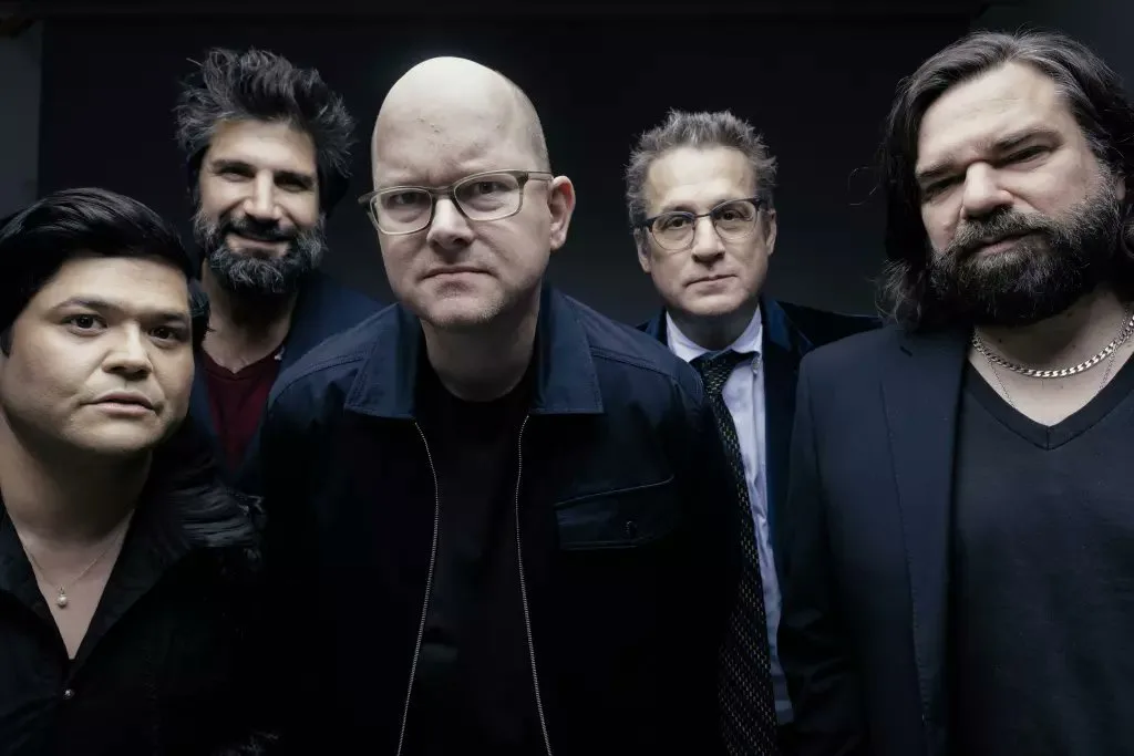A group photo of five men, mostly in dark clothing, with serious or deadpan expressions. The man in the center, bald and…