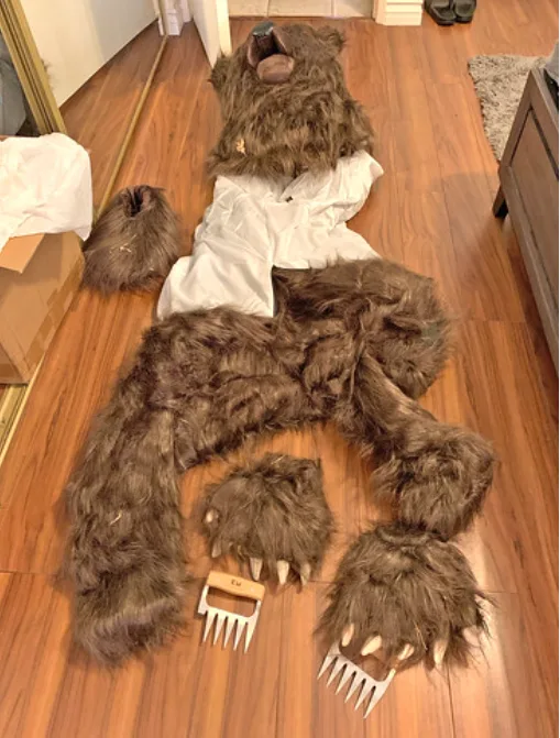 A large, brown, furry bear costume is laid out on a wooden floor, with its head, arms, legs, and paws separated and spread…