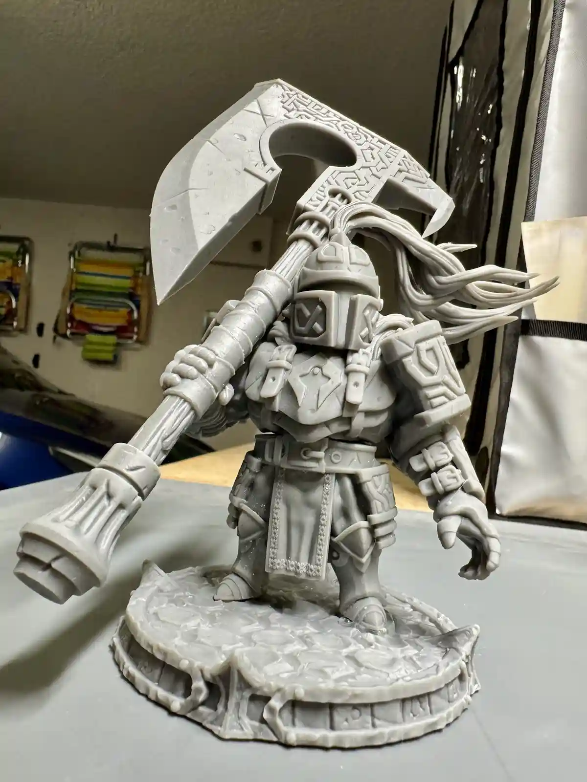 A gray, unpainted 3d-printed fantasy warrior figurine stands on a circular base, holding a massive, ornate axe over its…