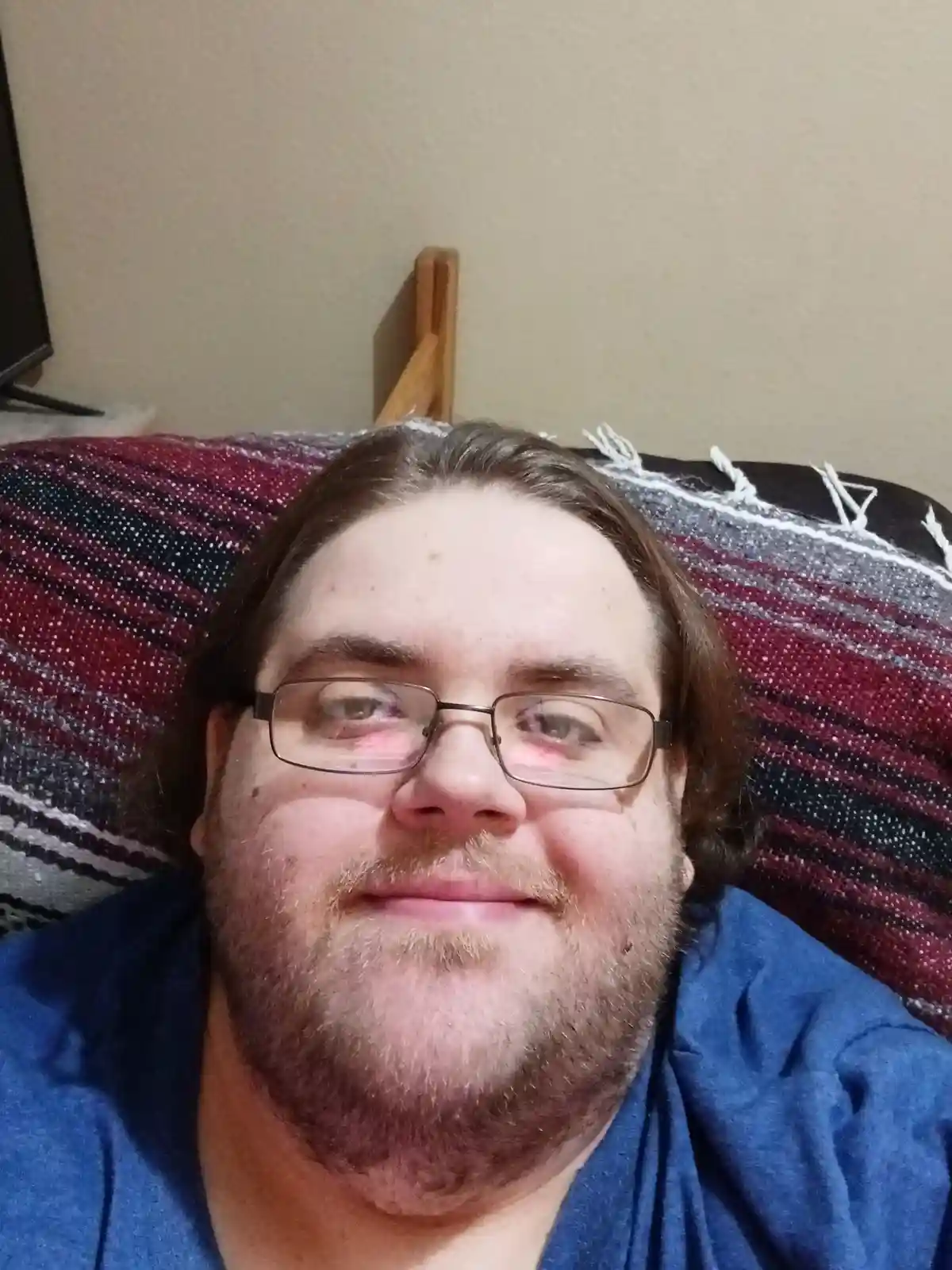 A man with glasses and a beard, wearing a blue shirt, lies on a striped blanket and smiles slightly while looking at the…