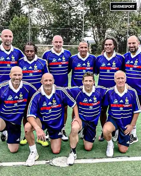 A group of older men in blue and white striped French football jerseys pose for a team photo on an artificial turf field…