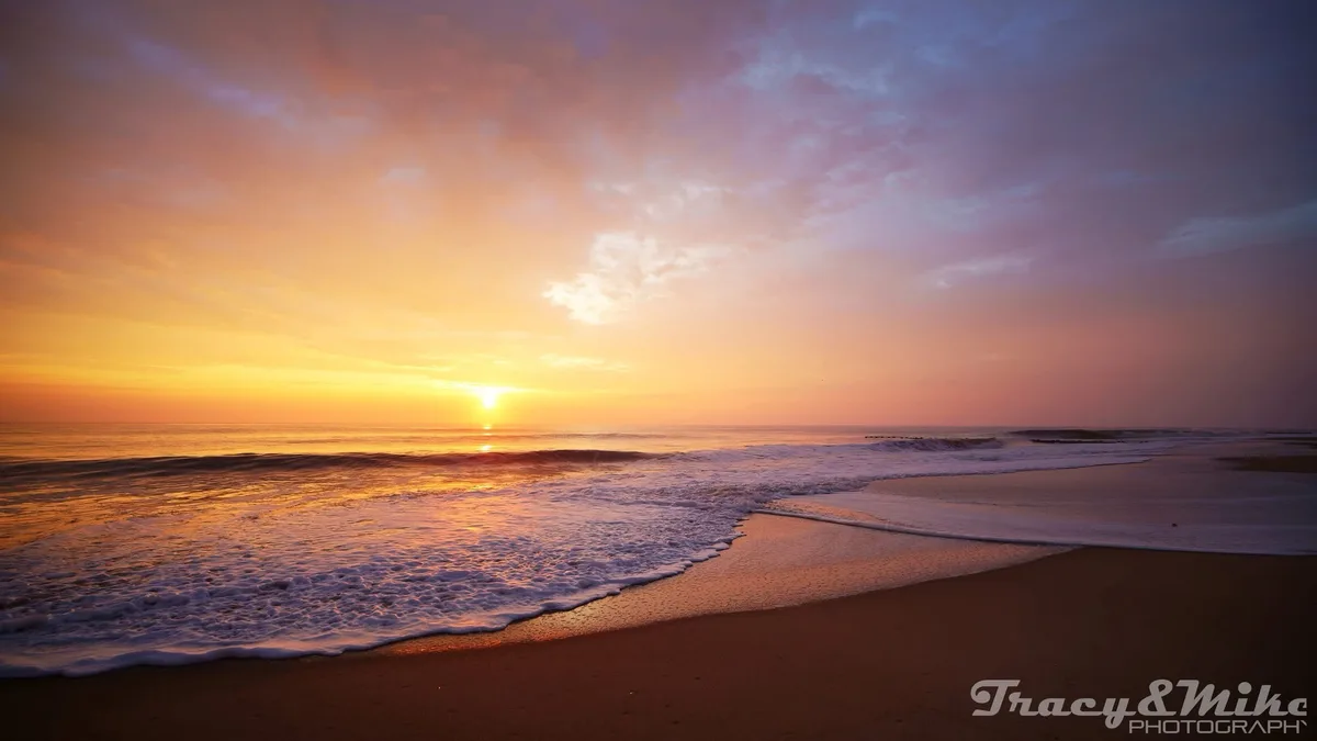 A serene beach sunset photo with the sun low on the horizon, casting orange and purple hues across the sky and reflecting on…