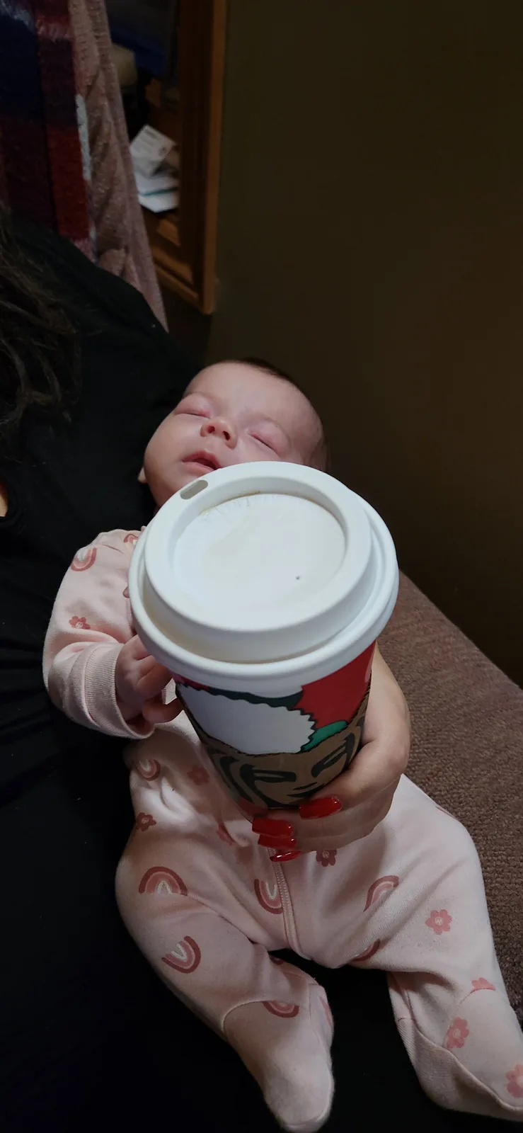 A sleeping baby in pink pajamas with rainbow and flower patterns is held by an adult and is holding a Starbucks coffee cup…