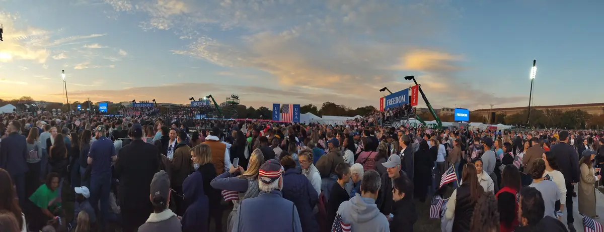 A wide-angle photo captures a large crowd gathered outdoors at dusk, many holding small American flags, facing a stage with…
