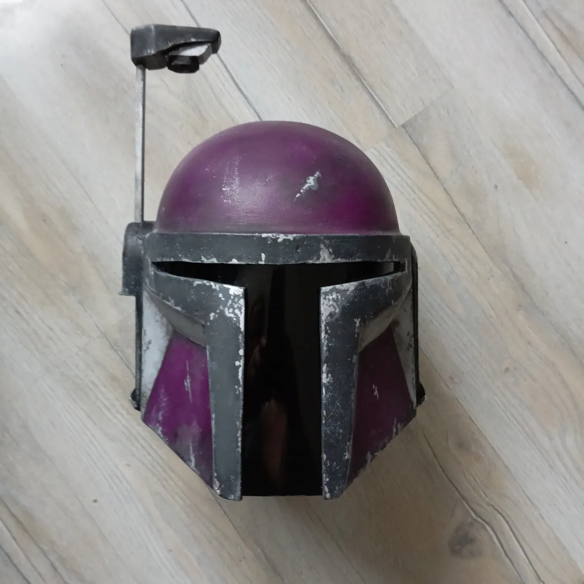 A purple and black Mandalorian-style helmet with a weathered, chipped finish sits on a light wood floor. The helmet has a…