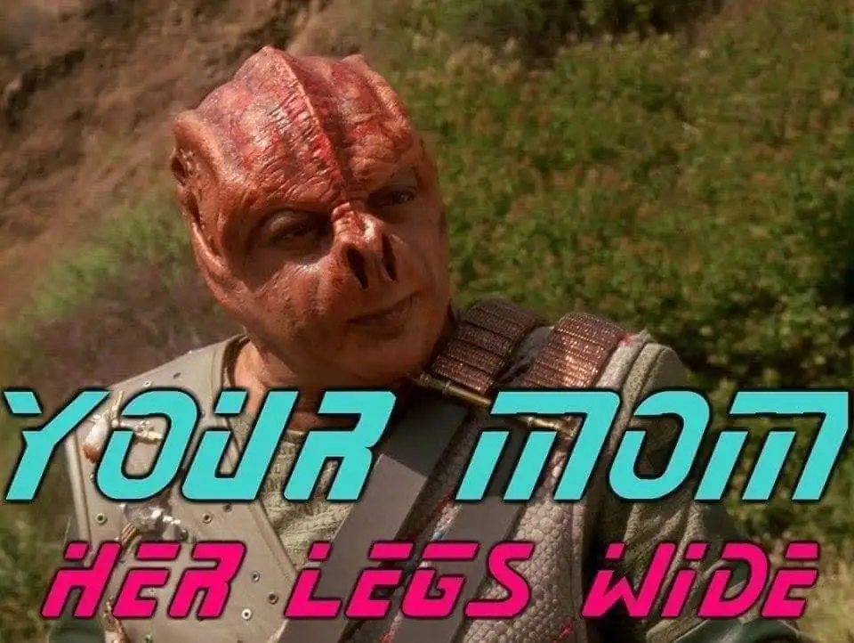 A close-up of a red-skinned, alien-looking man with a wrinkled face and large nose, wearing a textured vest, standing…