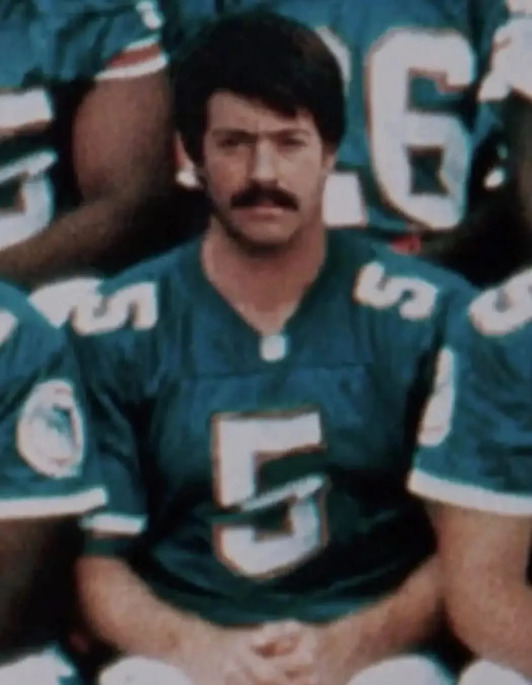 A vintage photograph shows a man with a mustache wearing a teal Miami Dolphins football jersey with the number 5. He is…