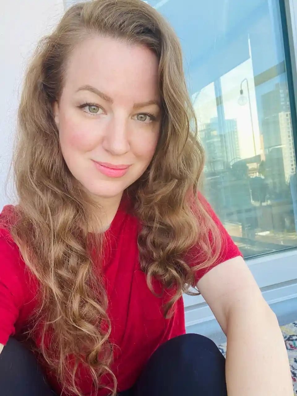 A woman with long, wavy brown hair and green eyes smiles gently at the camera while wearing a red t-shirt and dark pants…