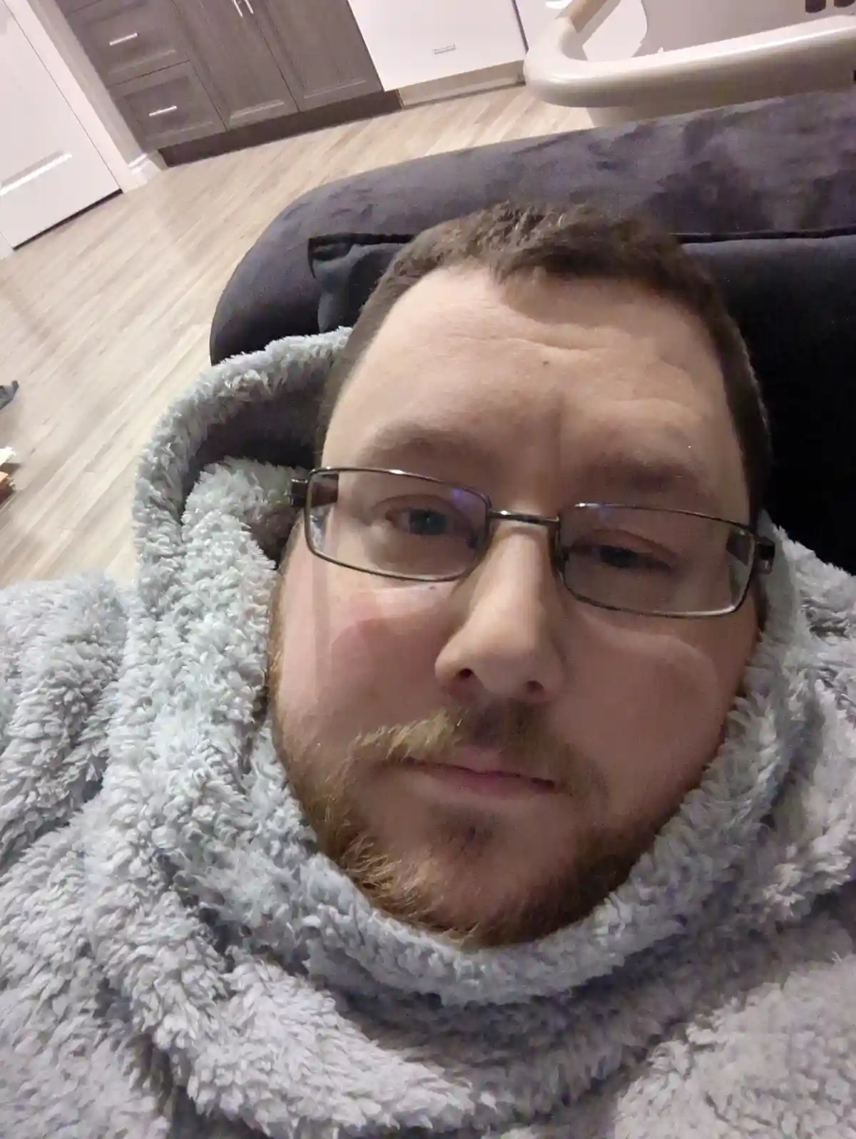 A man with glasses and a beard is lying down, wrapped in a fluffy grey hooded blanket, looking directly at the camera with a…