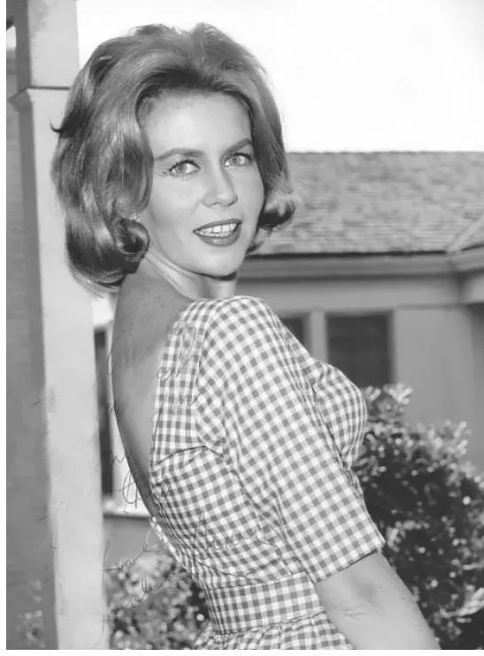 A black-and-white photo of a woman with styled hair, wearing a checkered dress, smiling while looking over her shoulder. The…
