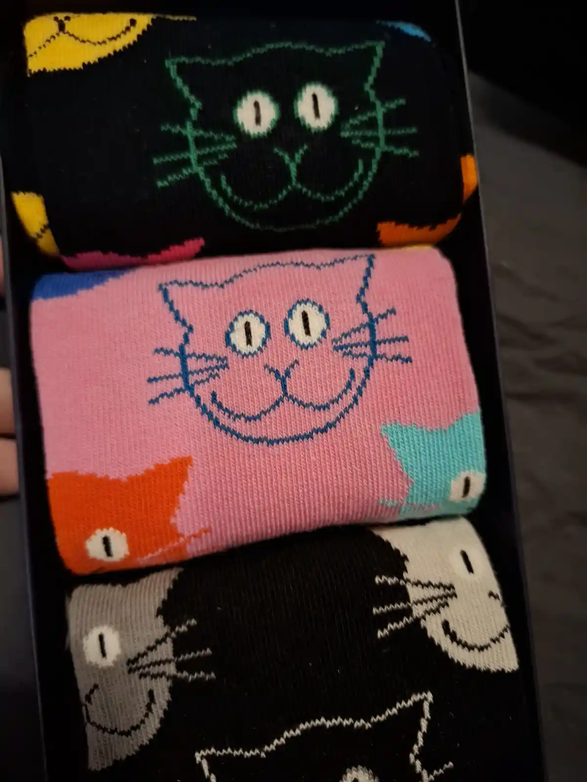 Three pairs of folded, colorful cat-themed socks are stacked in a box, each featuring a different cartoon cat face with wide…