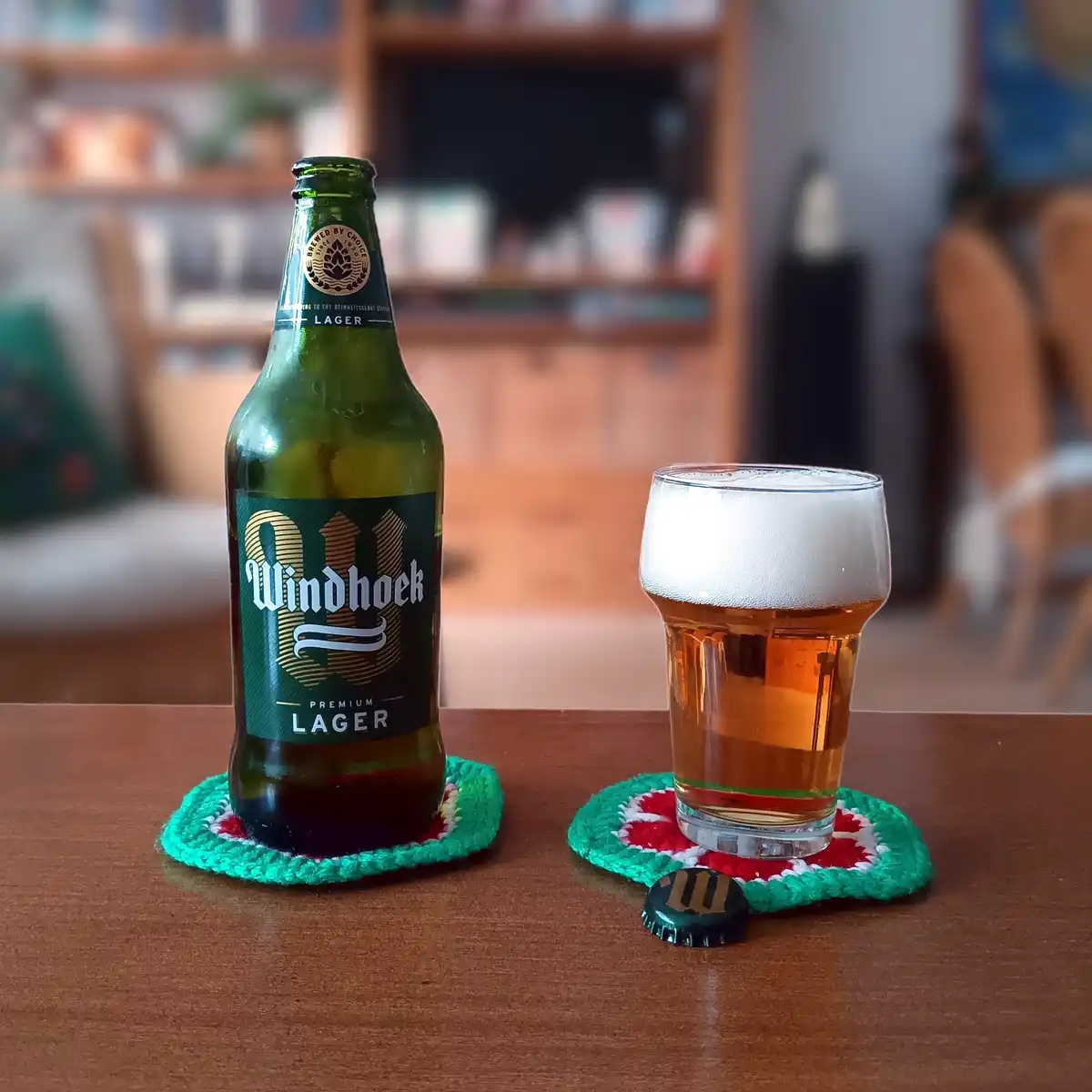 A green bottle of Windhoek Premium Lager sits next to a full glass of beer on matching crocheted coasters on a wooden table…