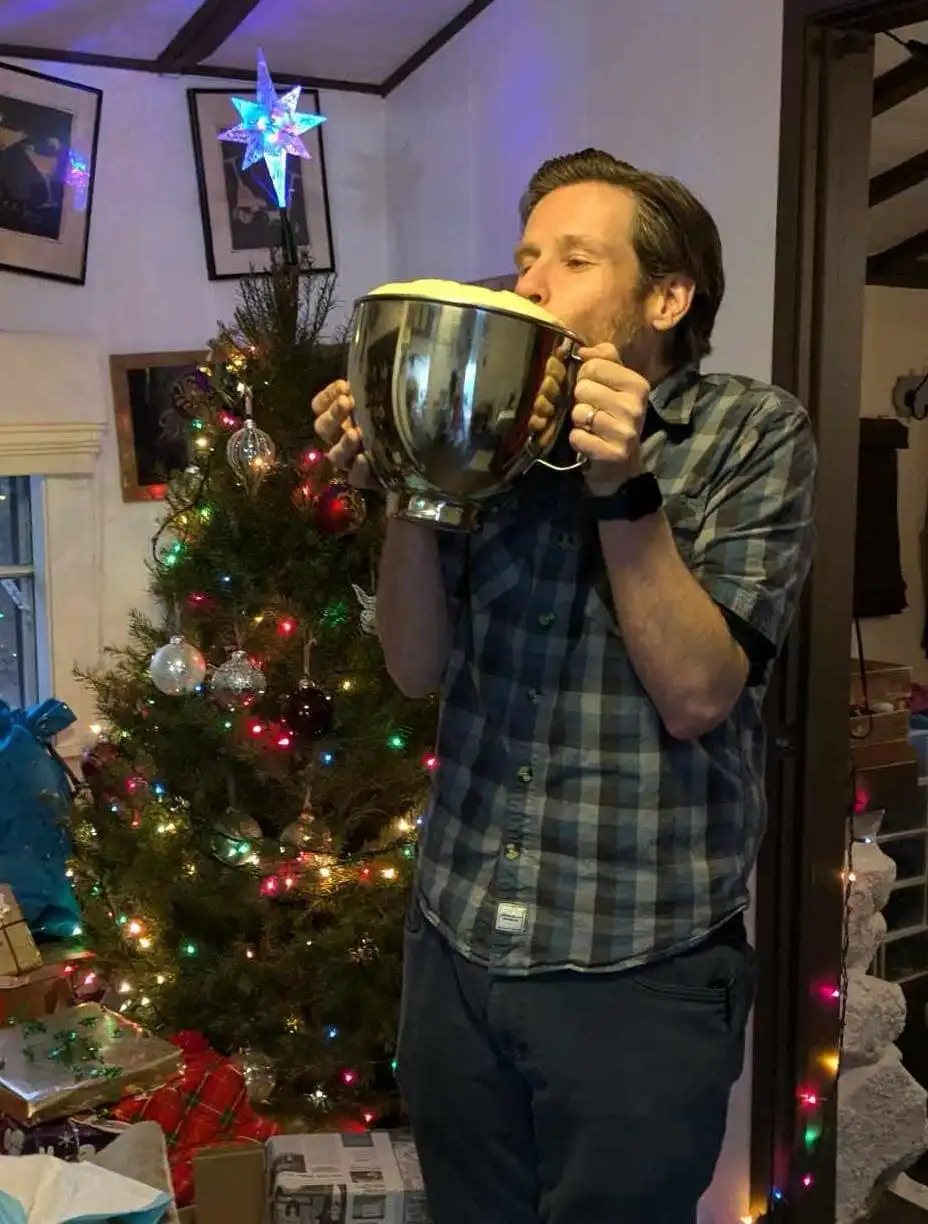 a man in a plaid shirt stands in a festively decorated room with a lit christmas tree behind him, holding a large metal…