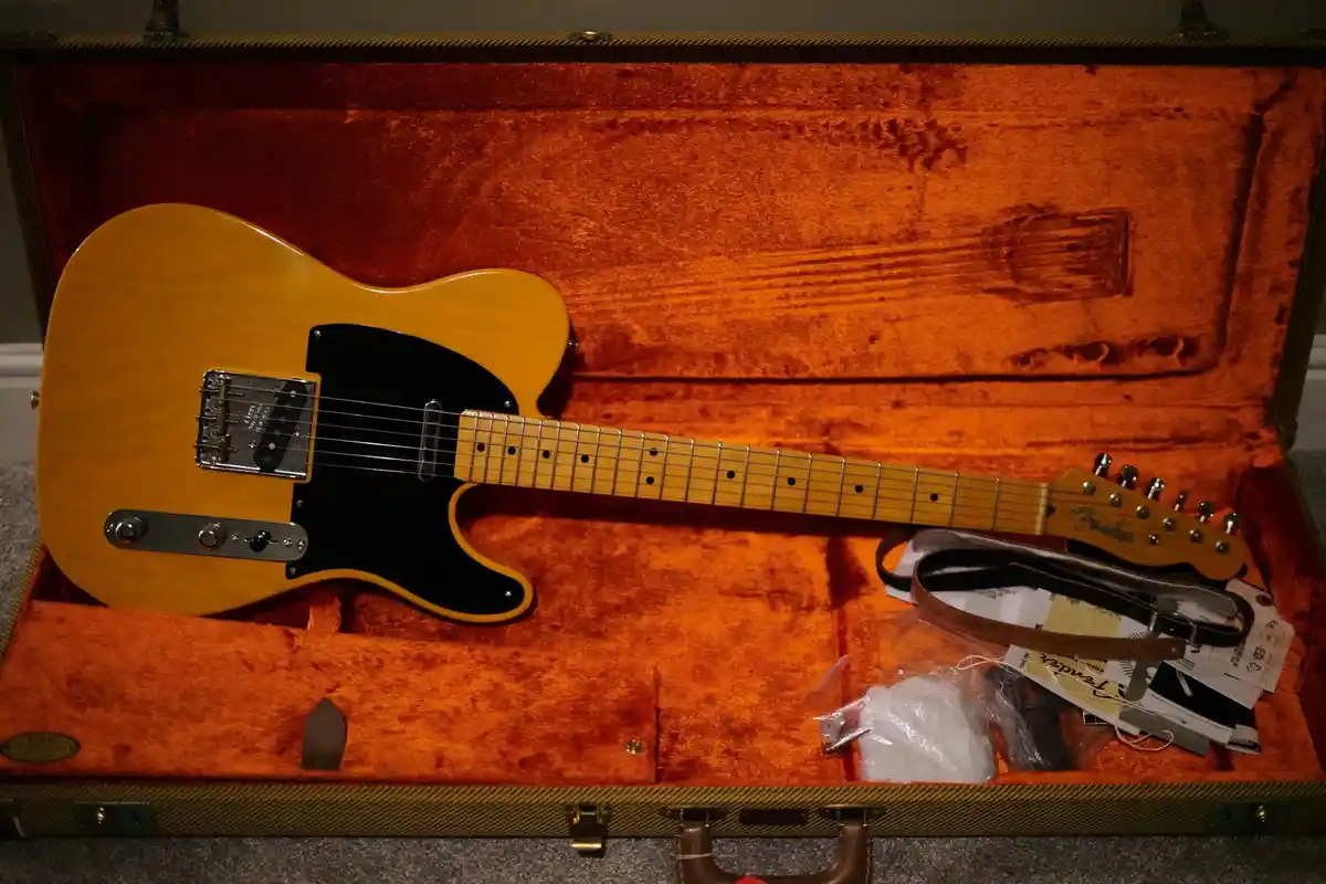 A light-wood Fender Telecaster electric guitar rests inside its open, orange velvet-lined hardshell case, with a brown…