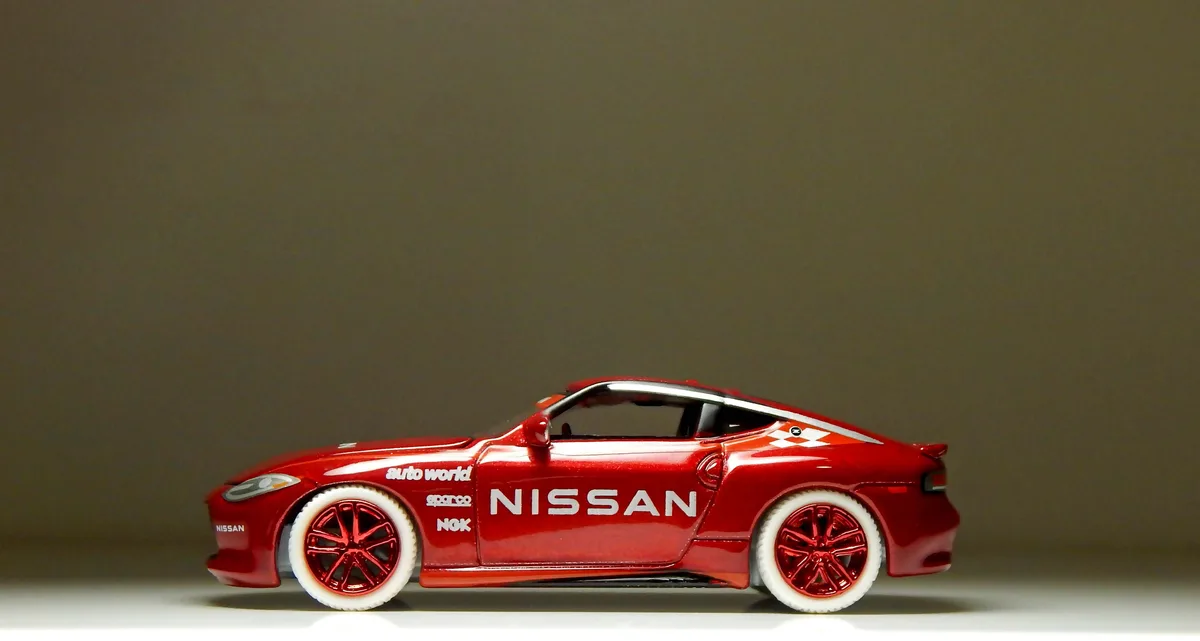 A red die-cast model of a Nissan sports car is shown in profile against a plain gray background. The car has white-wall…