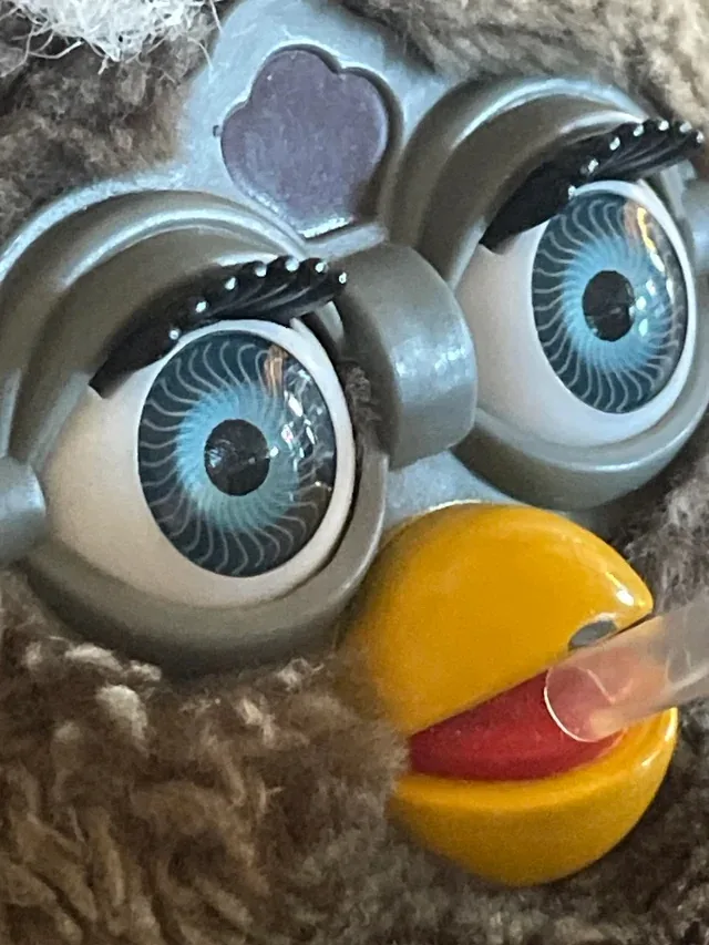 A close-up of a plush toy bird with large, detailed blue eyes and a yellow beak, where a human finger is visibly inserted…