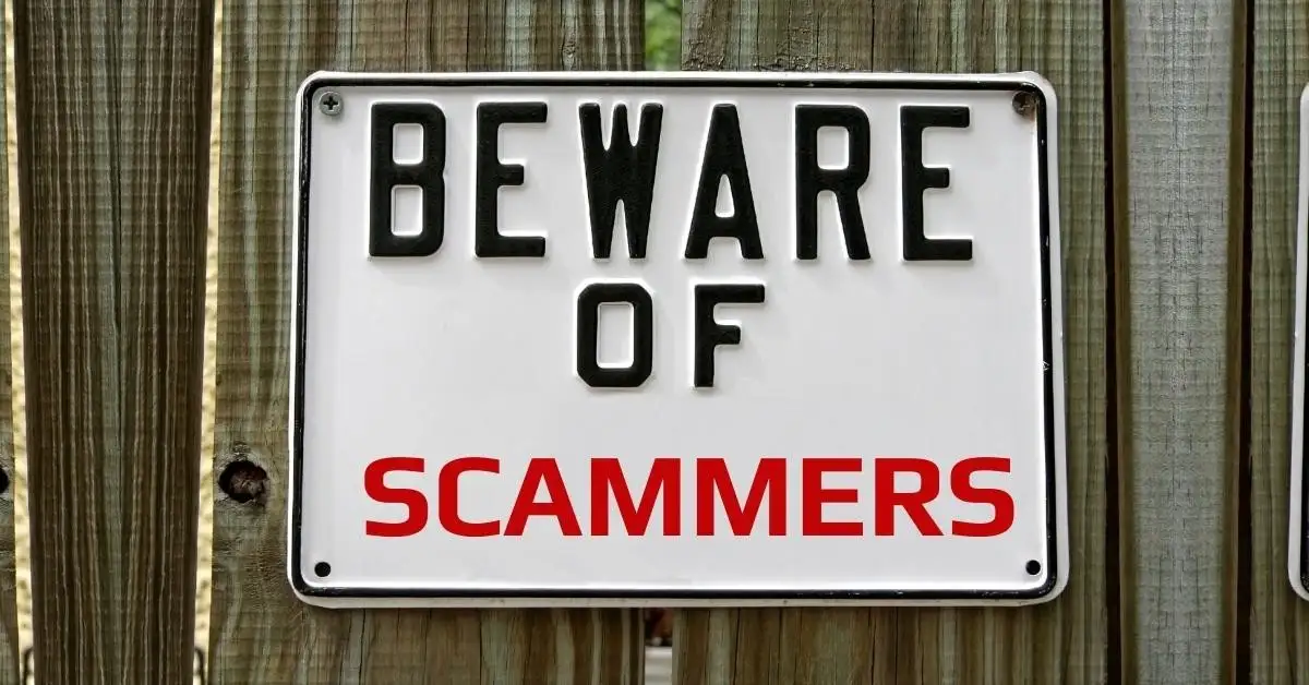A white rectangular warning sign with black and red lettering is mounted on a weathered wooden fence. The sign reads 'BEWARE…