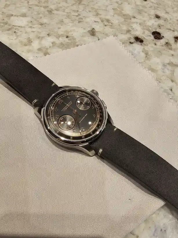 A close-up photo shows a luxury chronograph watch with a black dial, gold accents, and a dark suede strap resting on a beige…