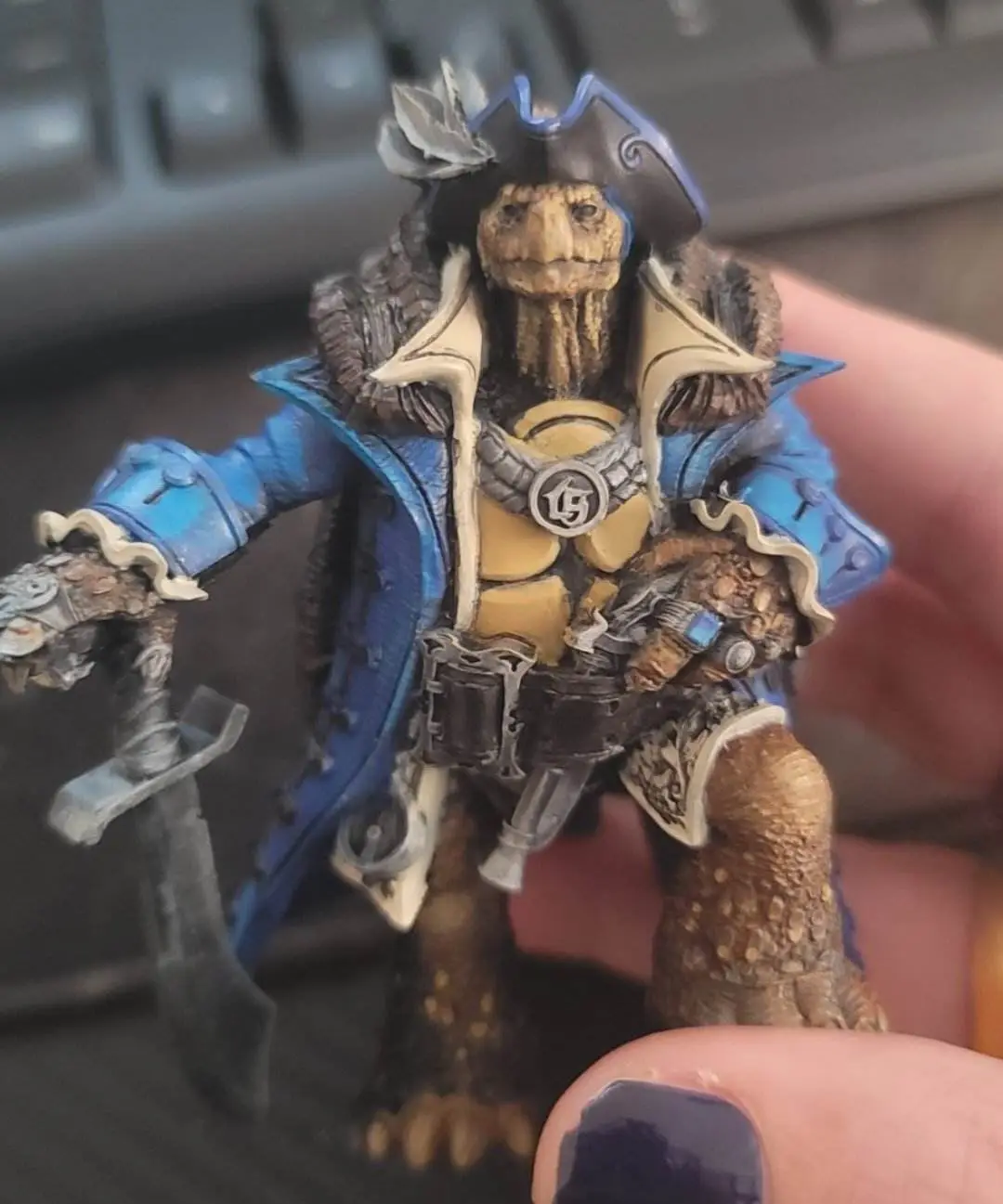 A hand holds a detailed miniature figurine of a turtle-like creature dressed as a pirate, wearing a tricorn hat, blue coat…