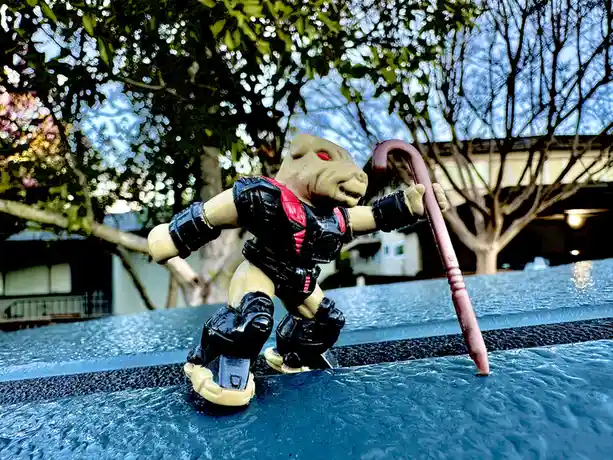 A small toy figure of a pig-like character in black and red armor stands on a textured blue surface, holding a pink cane…