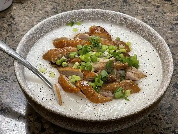 A close-up photo shows a bowl of congee topped with sliced roasted duck and chopped green onions, with a spoon resting in…