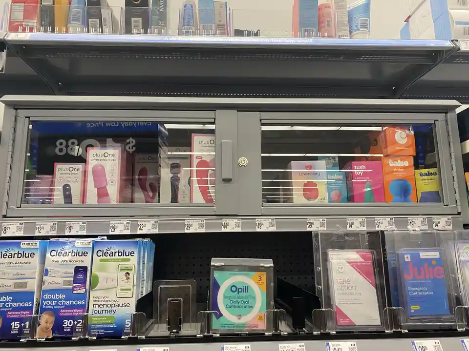 A store shelf displays personal care items, with a locked glass cabinet on the top shelf holding vibrators and other…