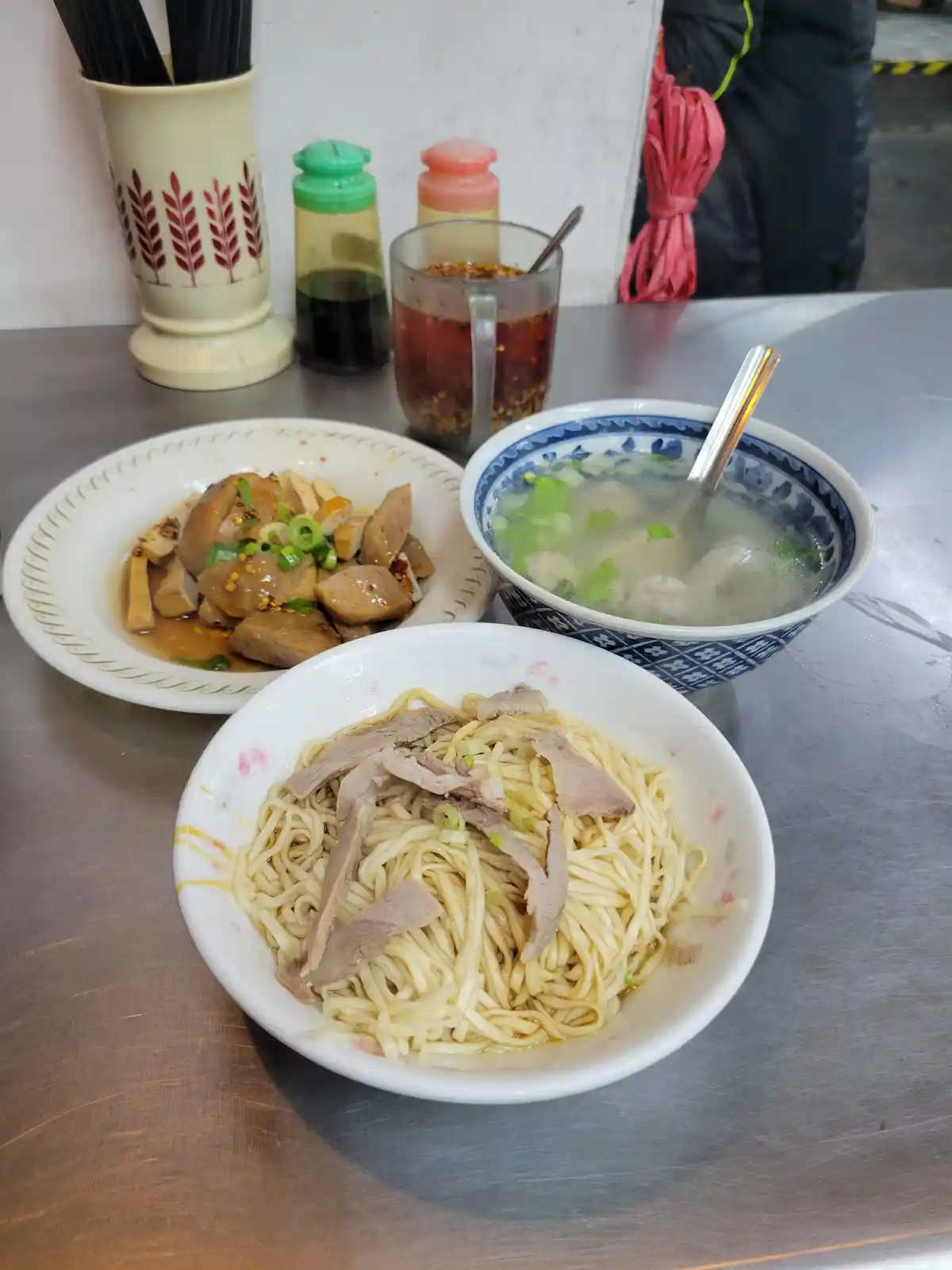 A table with three dishes: a bowl of noodles topped with sliced meat and green onions, a bowl of clear soup with meatballs…