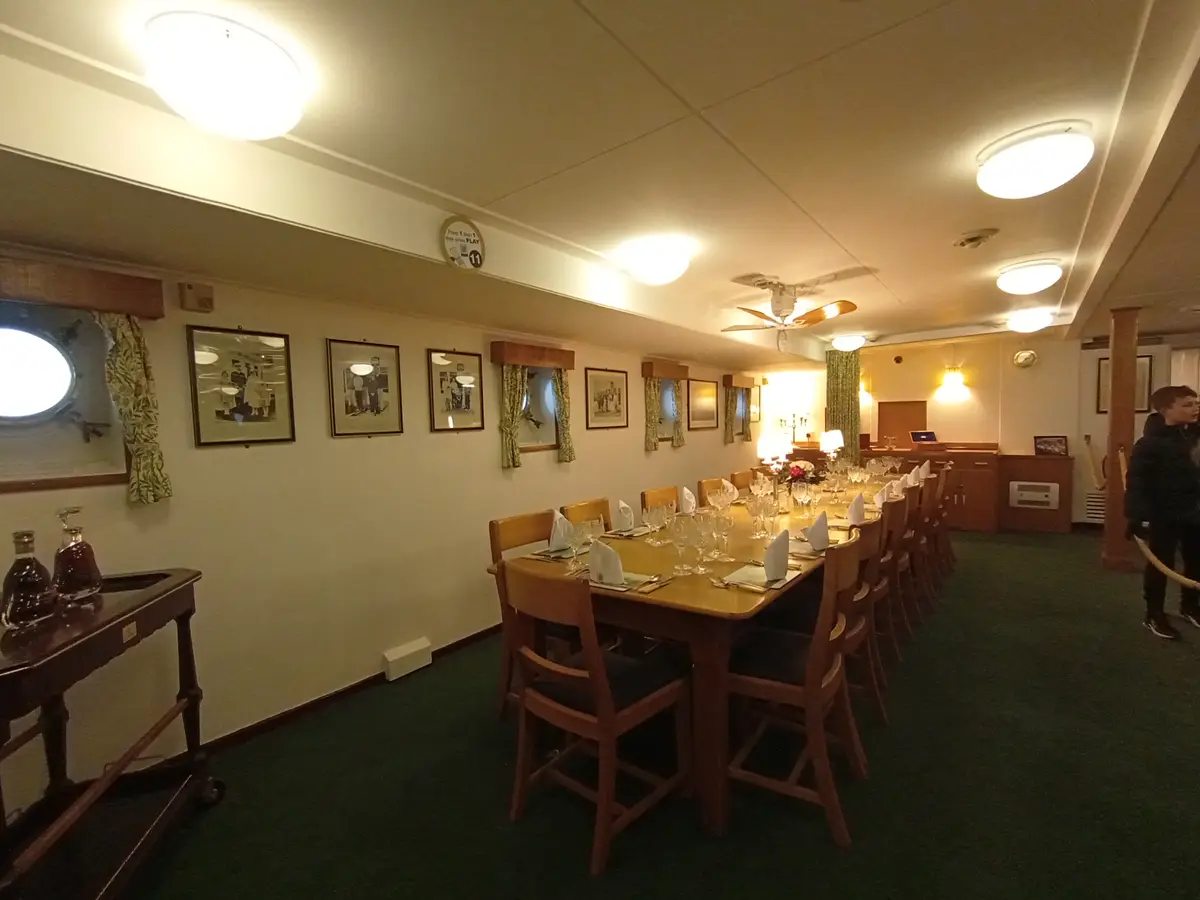 An empty, elegantly set dining room on a ship with a long wooden table, chairs, framed photos, and portholes, overlaid with…