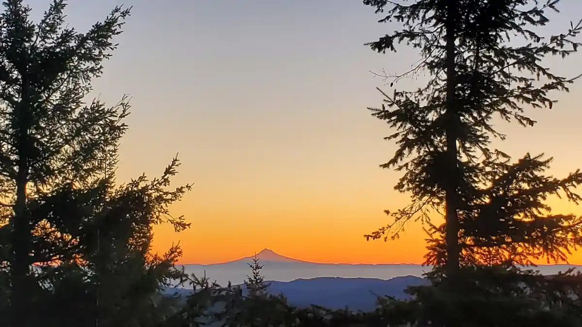 A scenic photo shows a mountain range at sunset, framed by the silhouettes of evergreen trees on both sides. The sky…