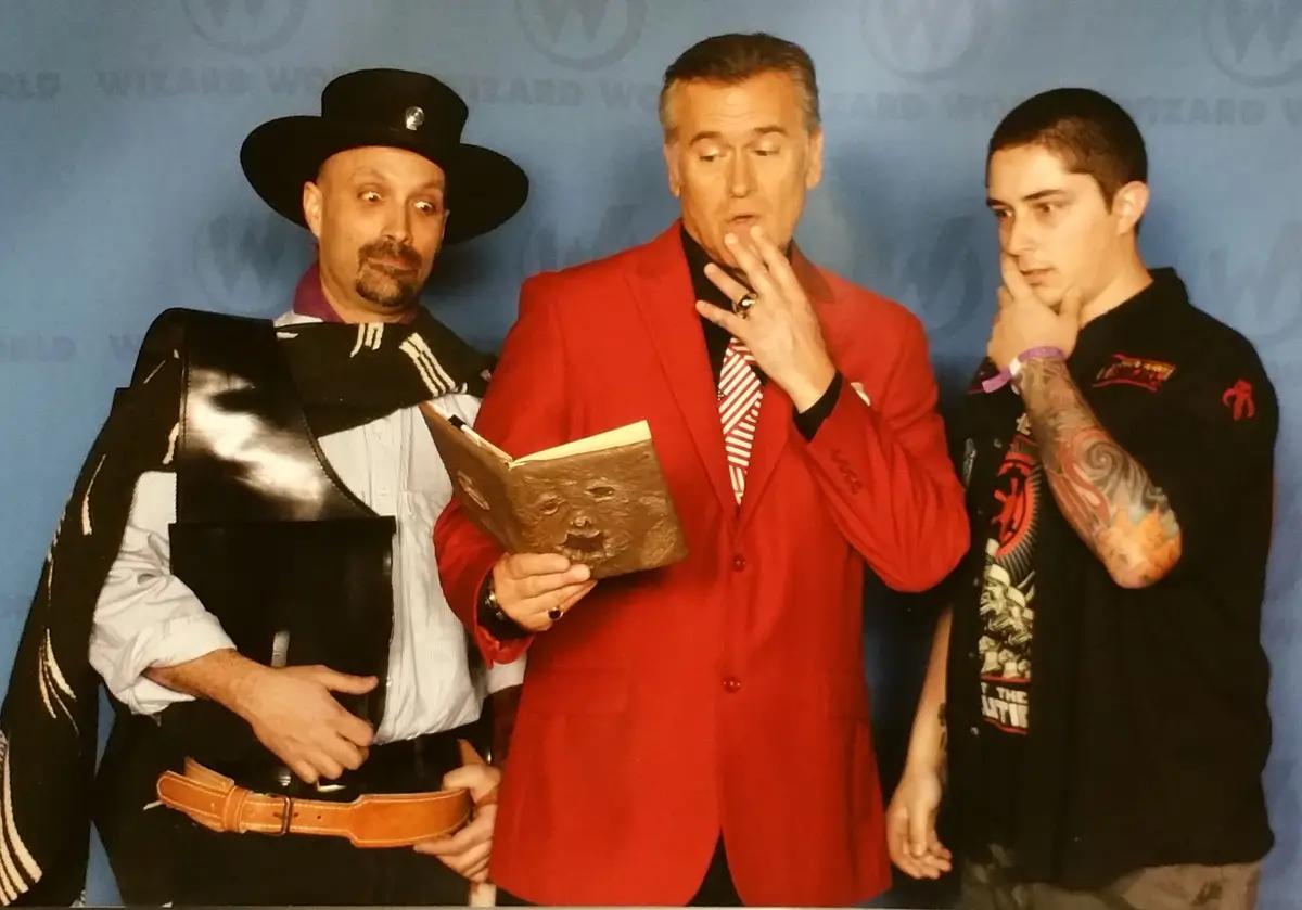Three men pose in front of a blue 'Wizard World' backdrop. The man on the left wears a black hat and shiny black vest. The…