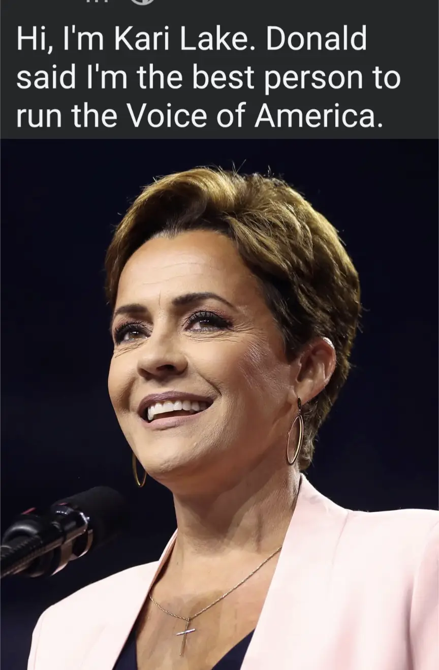 A close-up photo of Kari Lake smiling while speaking into a microphone, wearing a pink blazer, a cross necklace, and hoop…