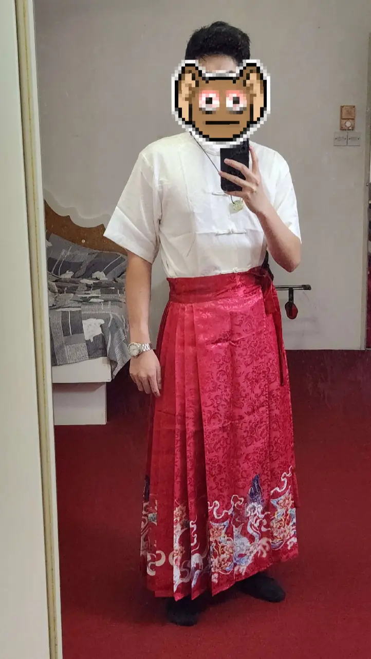 A person stands in front of a mirror wearing a white short-sleeved shirt and a long, ornate red skirt with traditional…