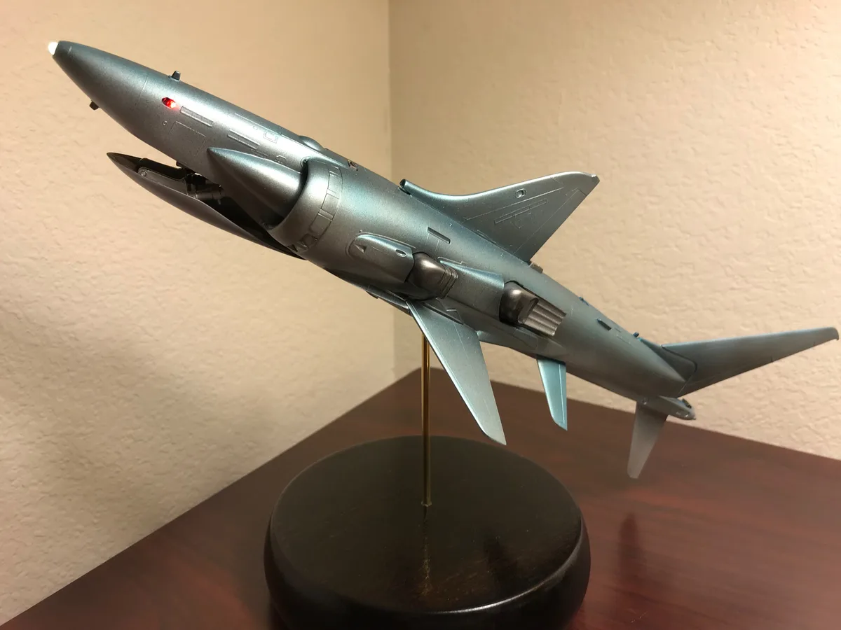 A detailed grey model airplane with a red light on its nose is mounted on a stand, angled upward. The overlaid text reads…