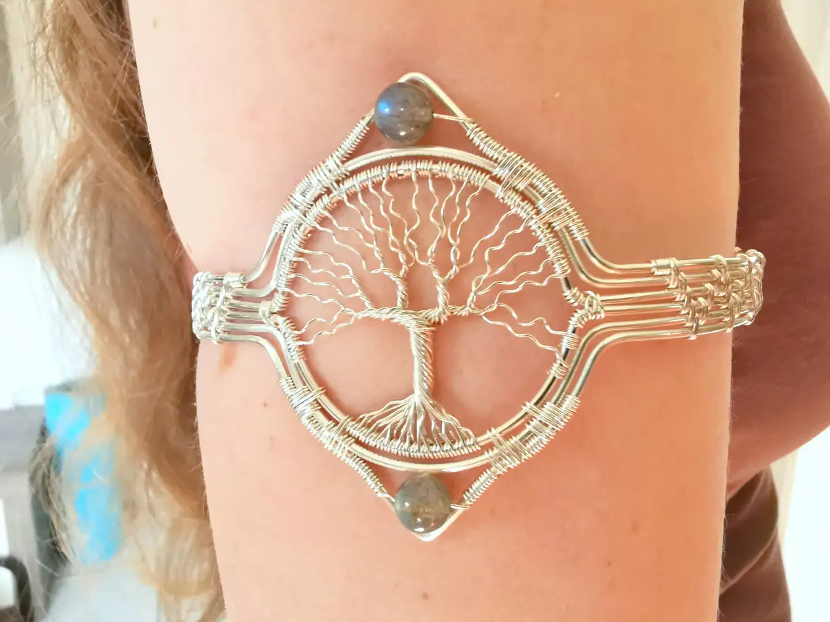 A close-up photo shows a person's arm wearing a large, intricate silver wire bracelet shaped like a tree of life with wavy…