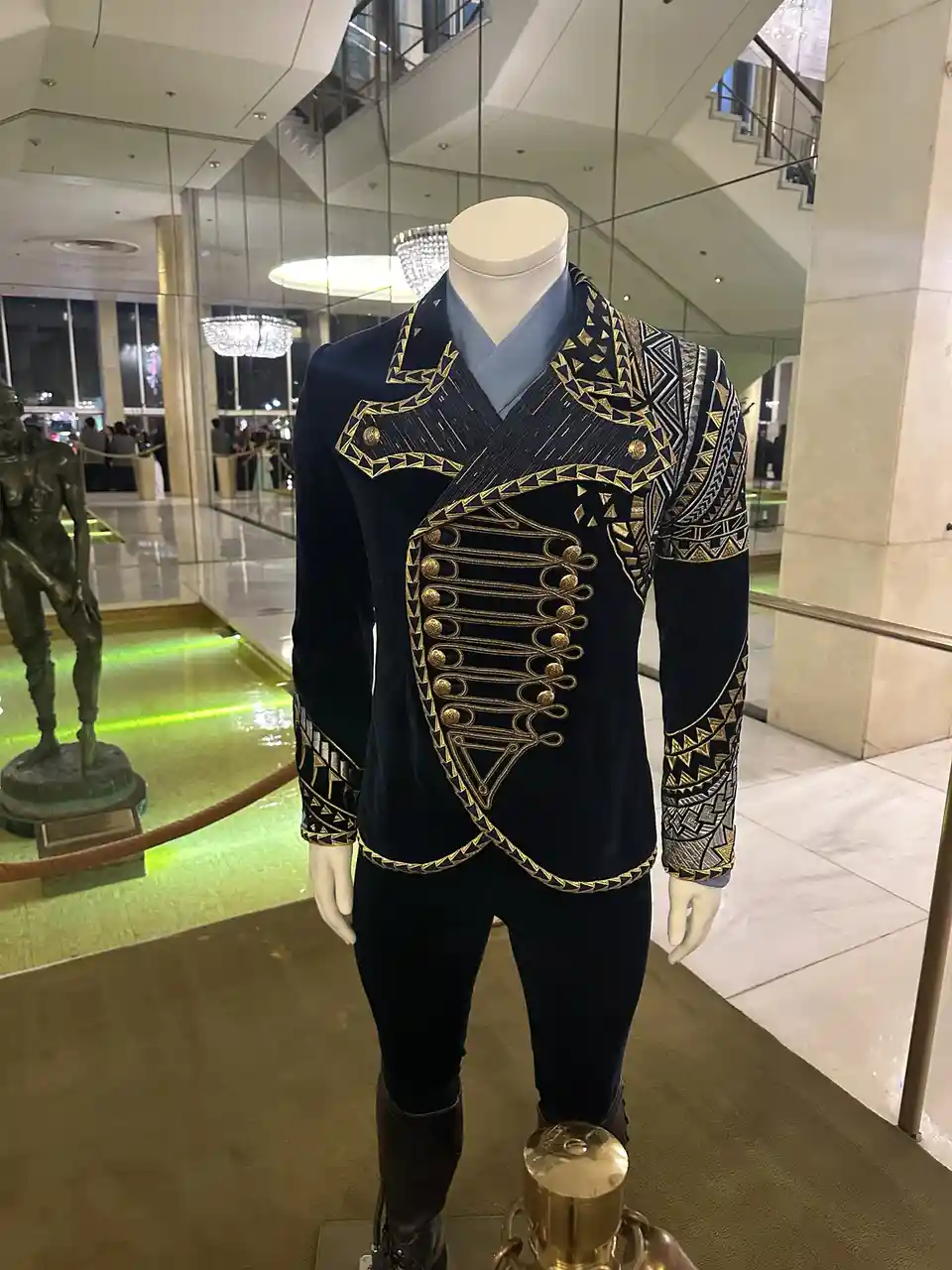 A mannequin dressed in an ornate, dark blue velvet jacket with gold geometric embroidery and gold buttons, paired with…