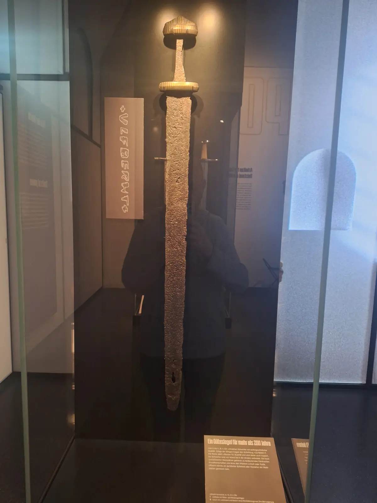 A large, ancient, rusted sword is displayed vertically inside a glass museum case, with the text '+ULFBERHT+' overlaid…