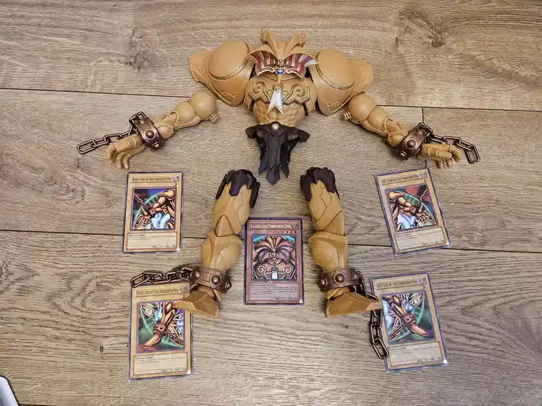 A tan-colored Exodia the Forbidden One action figure is laid out on a wooden floor, with its four limbs (two arms and two…