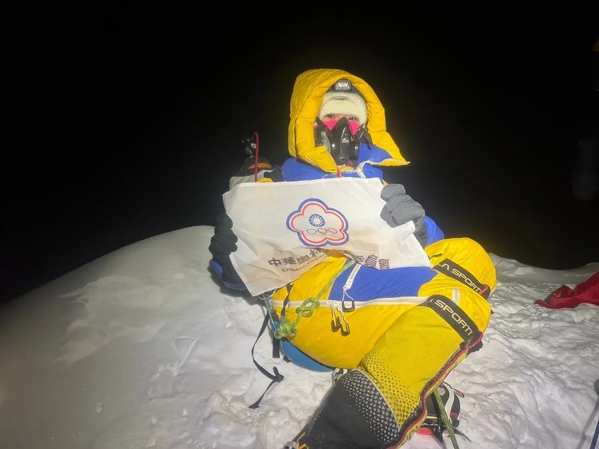 a person in full winter mountaineering gear, including a yellow hooded jacket, blue pants, and a pink face mask, sits atop a…