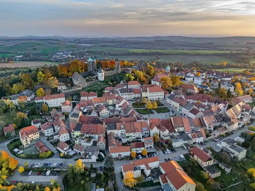 an aerial photograph of a picturesque european town with red-tiled roofs, stone buildings, and a castle on a hill surrounded…