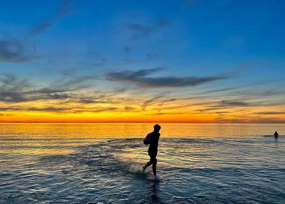 A silhouette of a person running through shallow ocean water at sunset, with vibrant orange and blue sky. Another person is…