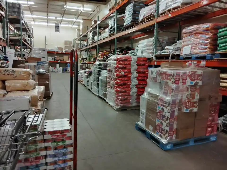 A wide-angle photo of a warehouse aisle filled with pallets of dog food and pet supplies, with a shopping cart in the…