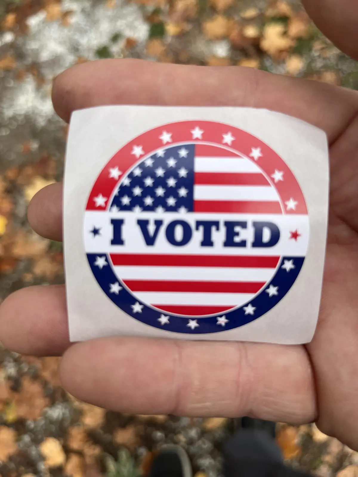 A hand holds up a circular 'I VOTED' sticker featuring American flag colors and stars, set against a blurred background of…