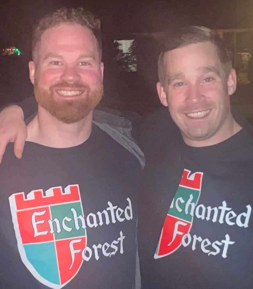 Two smiling men pose together at night, both wearing dark t-shirts with the 'Enchanted Forest' logo. The man on the left…