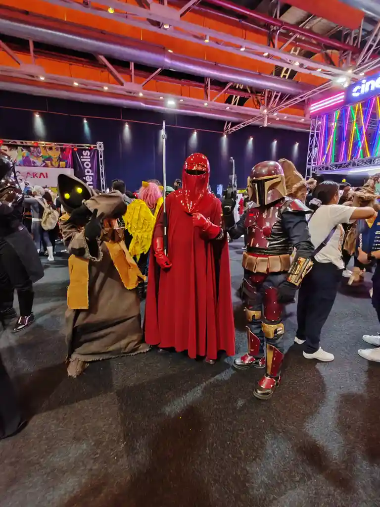 Three cosplayers at a convention: a character in a brown robe with glowing yellow eyes, a red hooded figure in a full red…