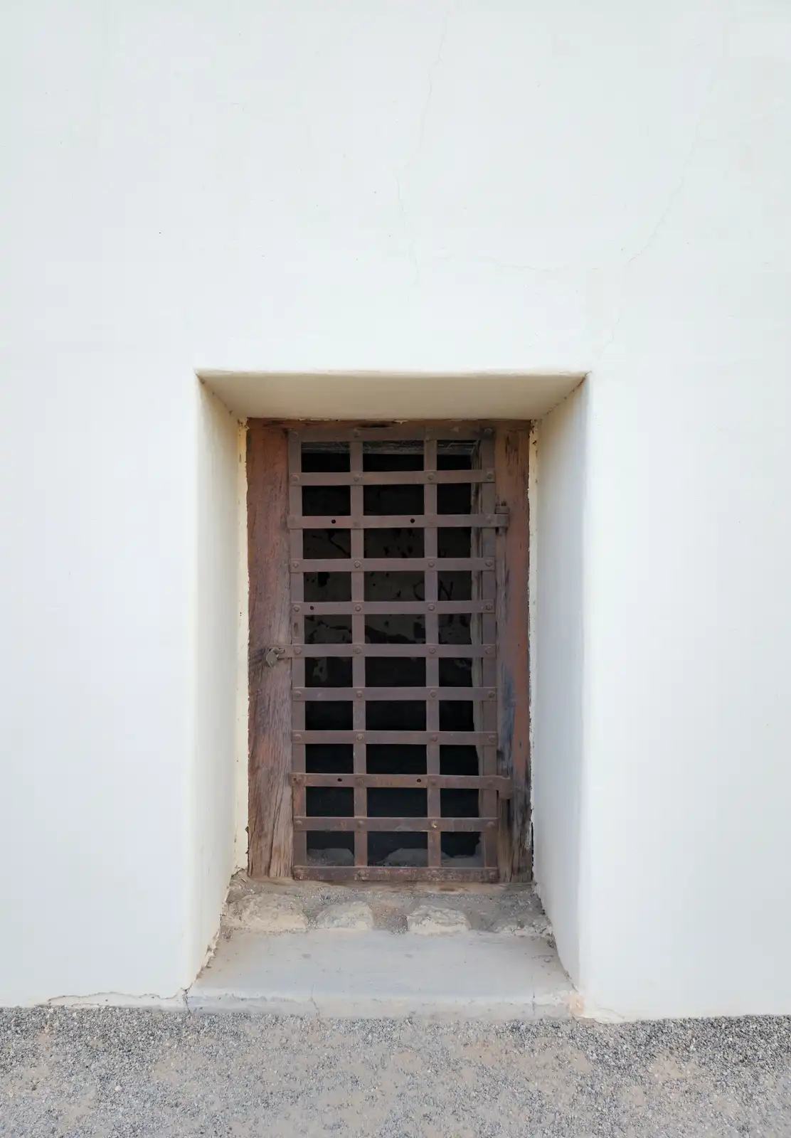 A small, barred wooden door set in a white stucco wall, with a rusty metal grate covering the opening. The overlaid text…