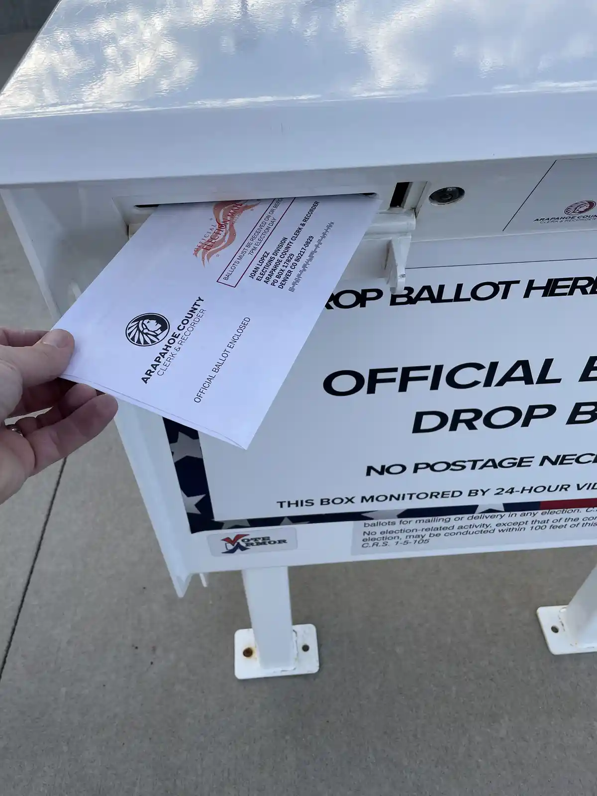 A hand holds an official Arapahoe County Clerk & Recorder ballot envelope partially inserted into a white official ballot…