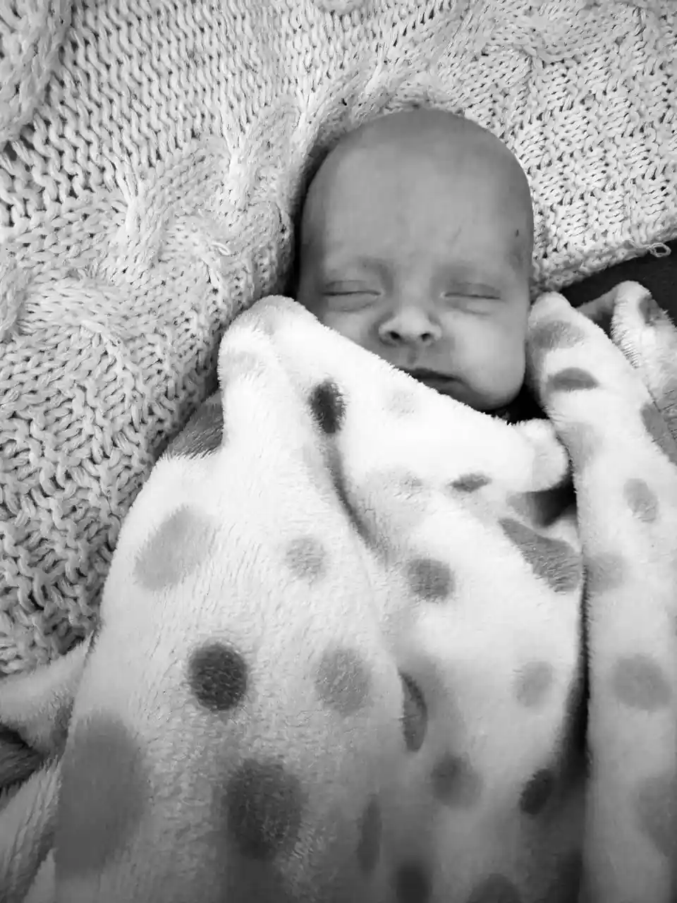 A black-and-white photo shows a sleeping newborn baby wrapped in a spotted blanket, resting on a textured knit blanket. The…