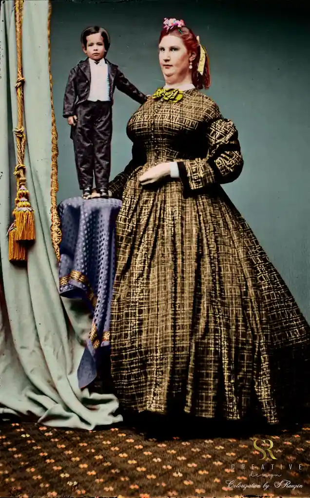 A colorized vintage photograph shows a woman in a large 19th-century dress standing beside a small boy in formal attire, who…