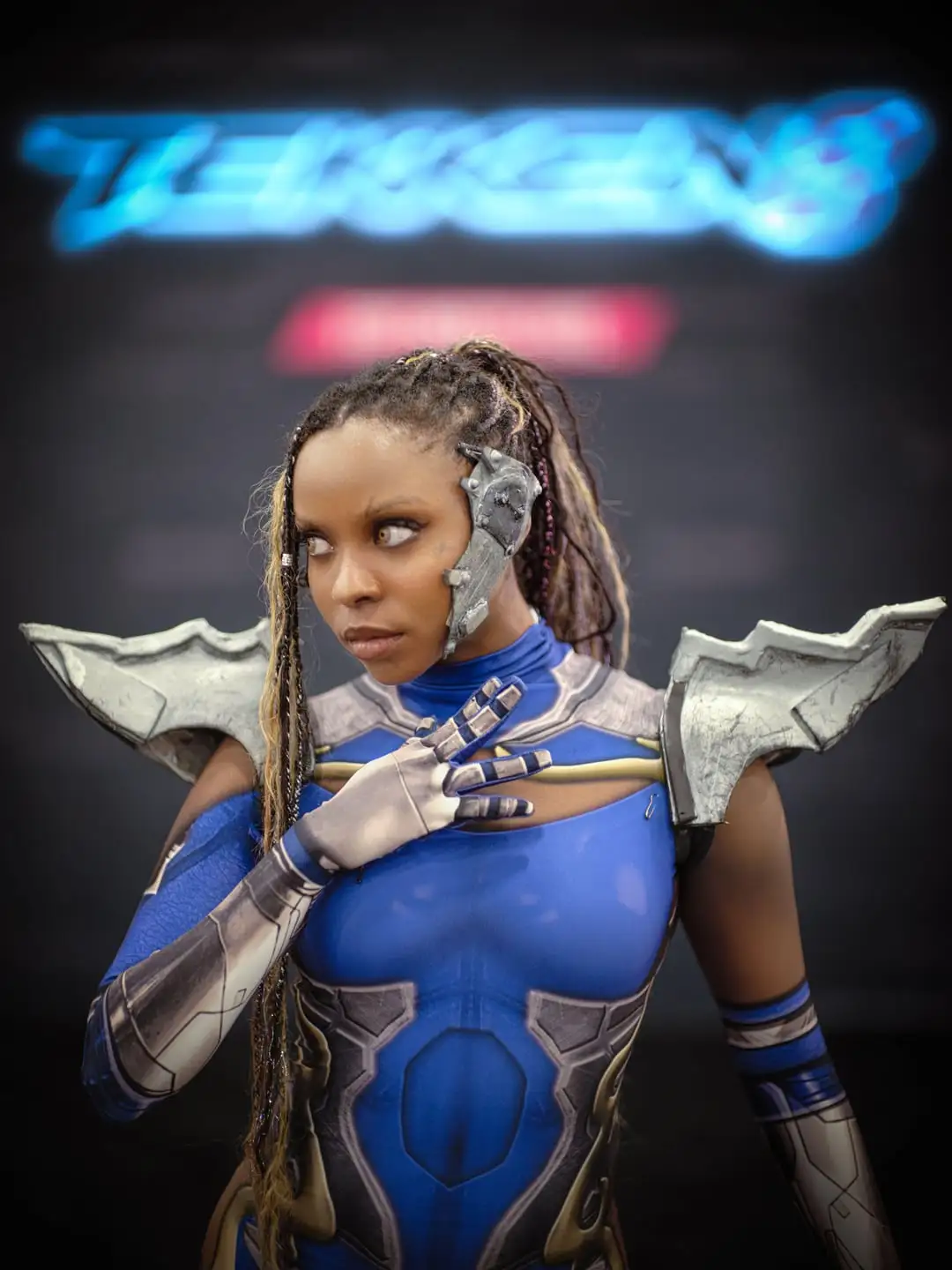 A cosplayer in a detailed blue and silver armored costume with mechanical shoulder pieces and a cybernetic earpiece, posing…
