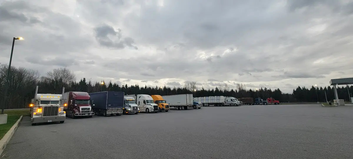 A wide shot of a large parking lot filled with numerous semi-trucks parked in a long line under an overcast sky, with trees…